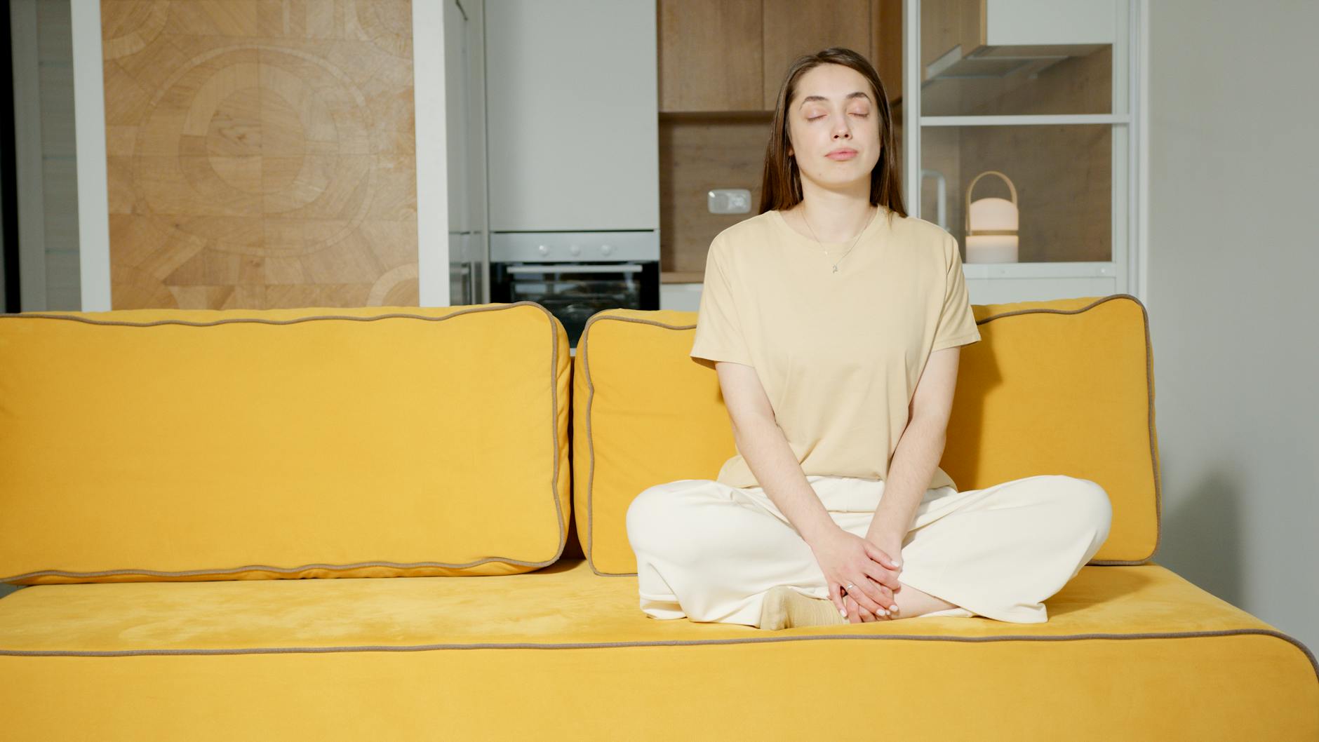 Relaxed woman meditating with closed eyes on a modern yellow couch in a minimalist home setting.
